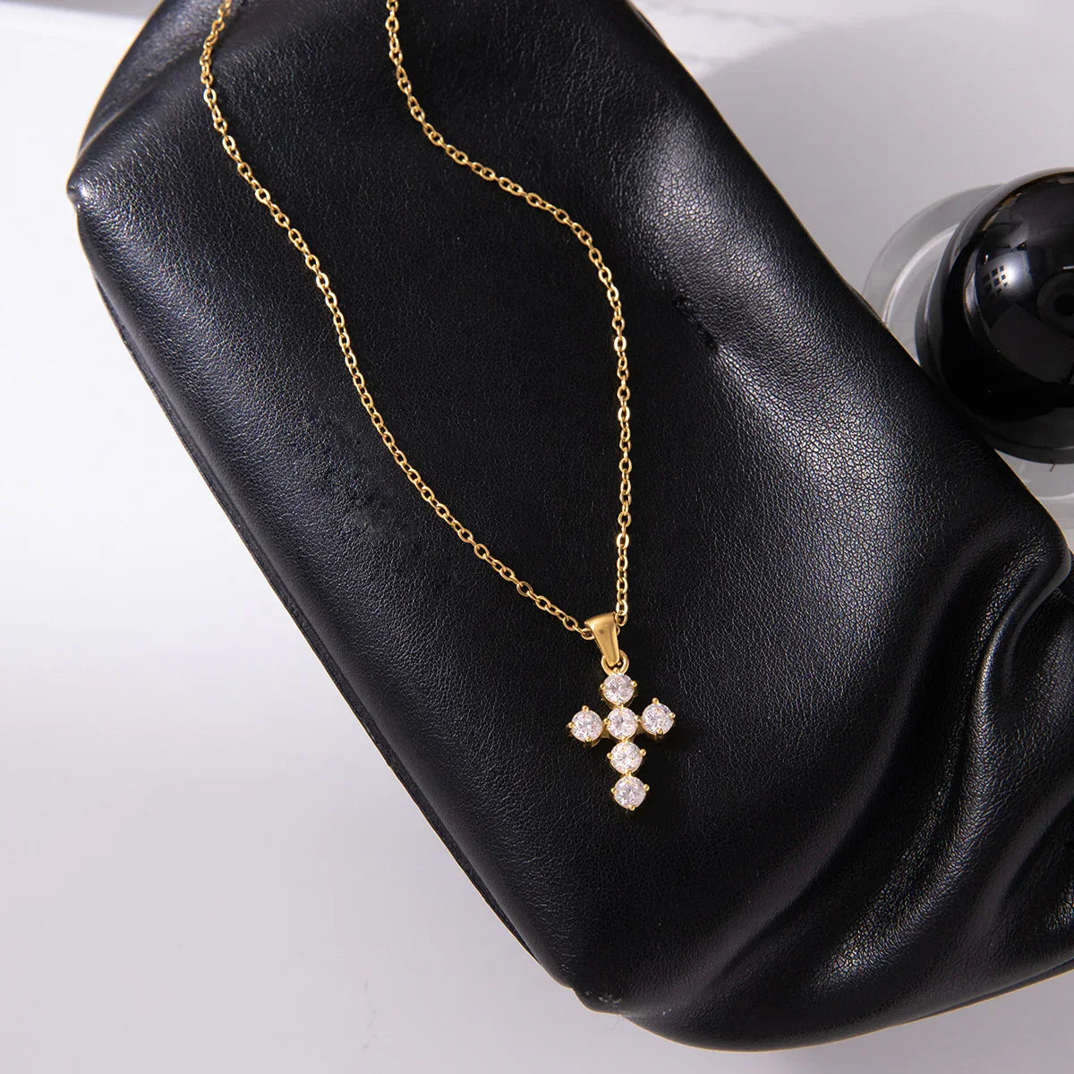 minimalist 18k gold cross pendant necklace close-up on black leather clutch background delicate jewelry detail