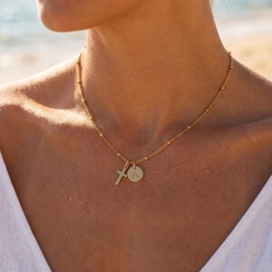 Woman wearing delicate 18K gold cross initial necklace at collarbone seaside outdoor portrait close-up