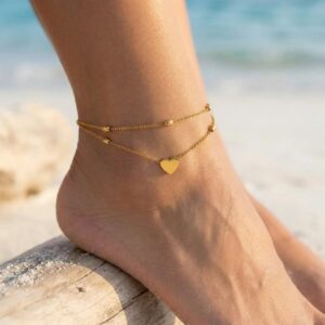 18K gold heart anklet worn on tanned female ankle at beach pier showing dainty dangling heart pendant and elegant layered chain design