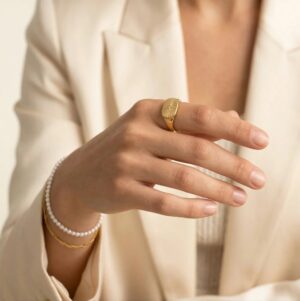 Gold signet ring worn on index finger, styled with beige blazer in professional studio setting showcasing elegant everyday wear