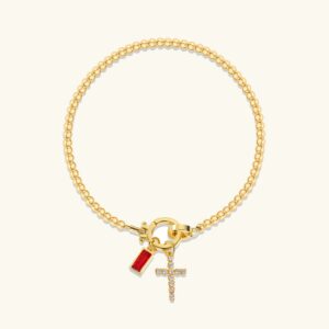 18K gold birthstone cross charm bracelet flat lay display showing beaded chain, pavé diamond cross, and red baguette-cut birthstone gem