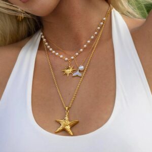 Layered 18K gold necklaces on model featuring starfish pendant, sun charm, mermaid tail charm and pearl chain detail worn on female model in white tank top