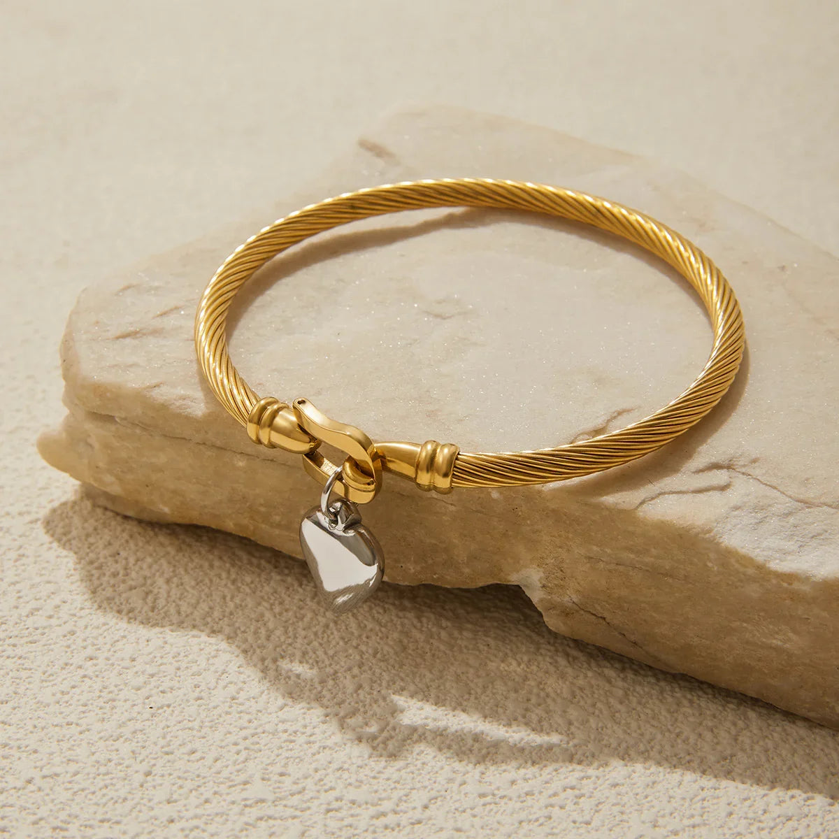 18K gold twisted cable bracelet front flat lay view on stone prop, polished twisted band with silver heart charm and secure clasp detail