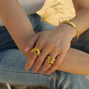 Gold heart ring worn on hand close-up with matching gold twist cuff bangle and band ring on model wearing denim and tank top