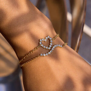 Layered 18K gold heart diamond bracelets worn on tanned wrist close-up showing dual delicate chains with pave crystal heart pendant charm in outdoor natural lighting