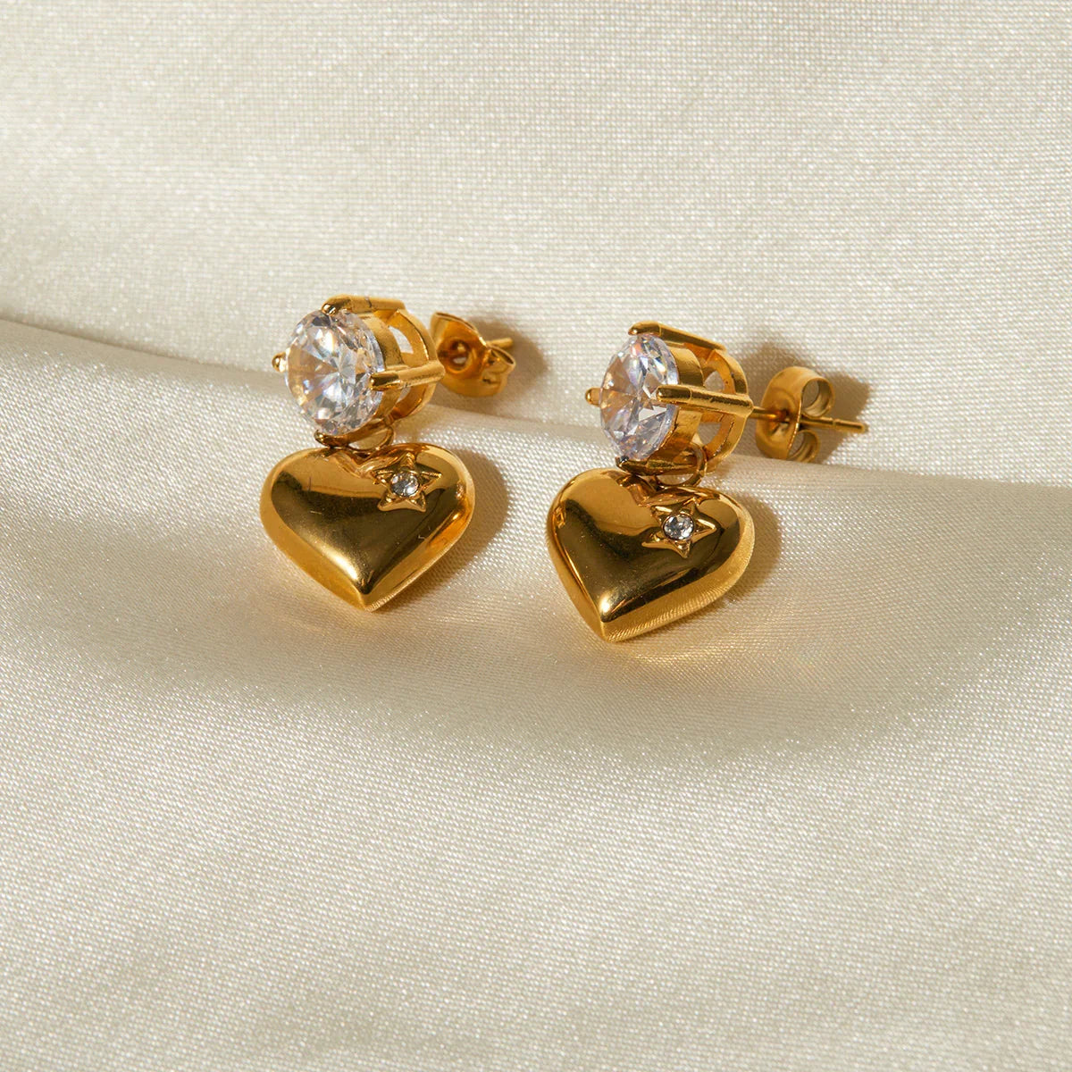 Pair of 18K gold heart drop earrings on cream silk satin fabric background, flat lay product photography showing shiny polished gold finish and cubic zirconia stones