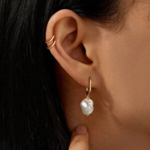 Woman wearing 18K gold pearl hoop earrings on ear with dark hair, close-up shot showing baroque pearl details and gold hoop design on pierced earlobe