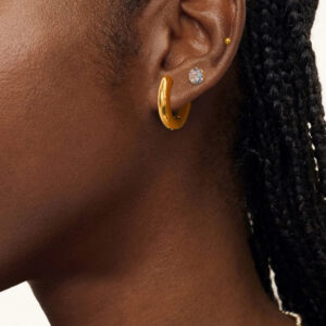 Close-up view of 18K gold earring bundle worn on model's ear, showcasing polished gold studs, cubic zirconia diamond studs, and huggie hoops with braided hair detail