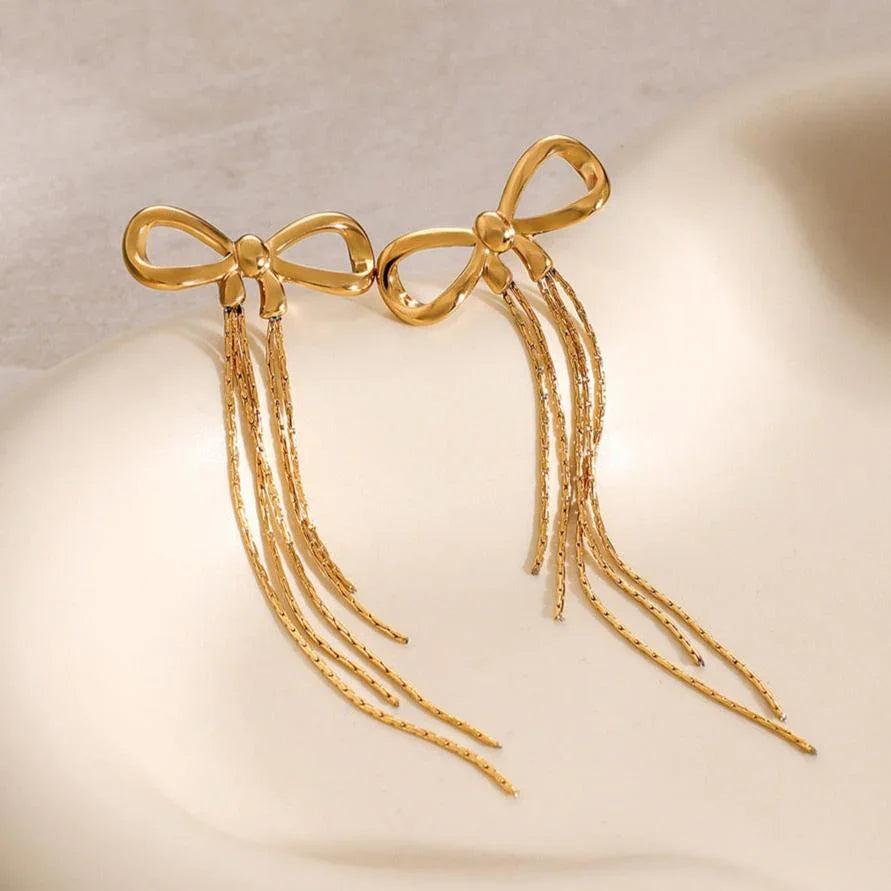 Matching pair of 18K gold bow tassel earrings on curved white surface, twisted chain design