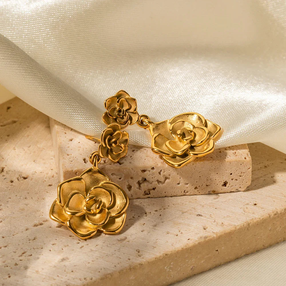 18K gold camellia drop earrings low angle flat lay on stone blocks and satin fabric showing carved layered flower petals matte golden texture