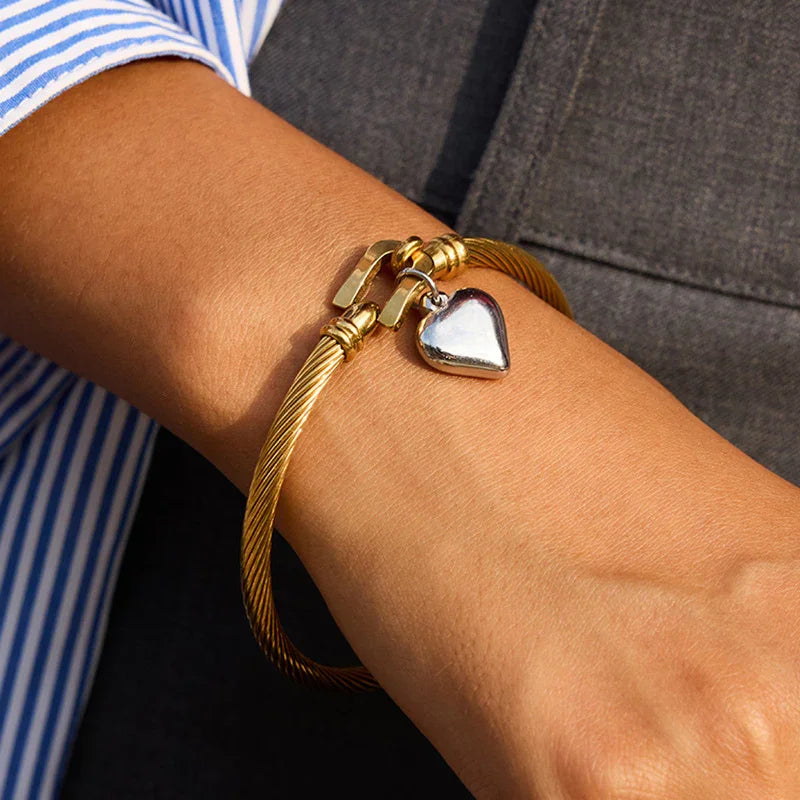 18K gold twisted cable bracelet worn on woman's wrist, close-up side view showing snug fit, hinged clasp, and dangling heart pendant in casual work setting