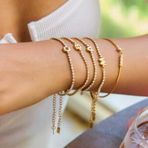 Stacked personalized initial tennis bracelets on female forearm, layered 18K gold charm design indoor