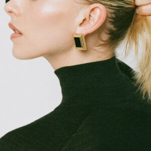 Close-up side profile of blonde woman wearing black and gold square enamel stud earrings on earlobe with half-up ponytail hairstyle
