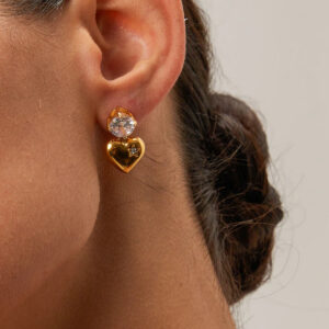 18K gold heart earrings worn on ear, close-up side view on model with low bun hairstyle, showing dangling heart charm with cubic zirconia accent stone