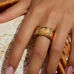 18K gold shell ring worn on female hand with light pink manicure, close-up detail view on vintage map background, showing polished gold tone and embossed seashell design