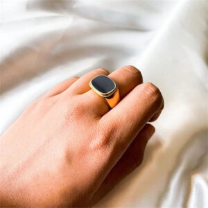 Gold signet ring with black enamel worn on male model's index finger against white satin fabric, close-up hand detail view showing polished glossy finish and band shape