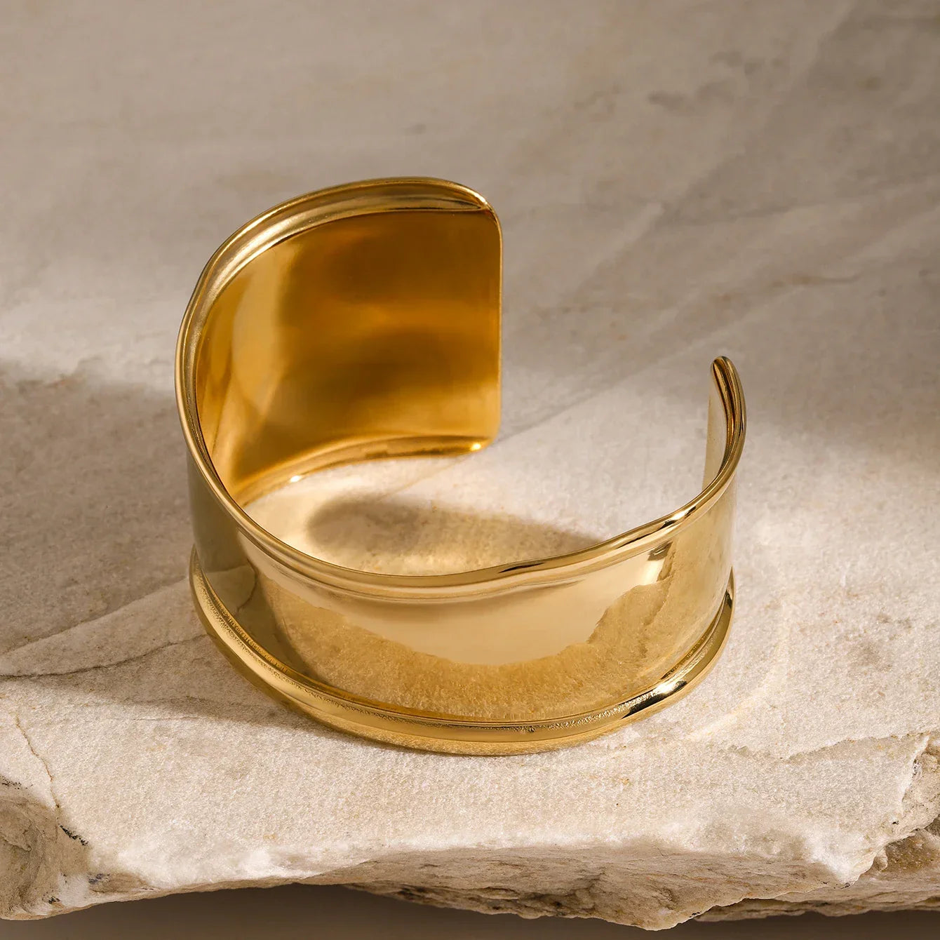 18K gold thick band cuff bracelet flat lay overhead, displaying highly polished reflective gold surface and substantial construction on stone slab background
