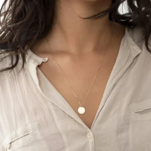 Minimalist gold disc pendant necklace worn by model, close-up upper neck view showing delicate thin chain and smooth round pendant with linen shirt