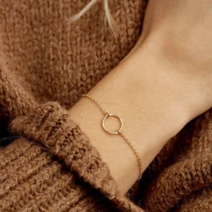 Model wearing delicate gold hoop charm bracelet on wrist with brown knit sweater - minimalist slim bracelet design in lifestyle setting