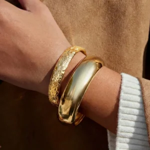 18K gold bangle duo stacked on wrist featuring polished smooth cuff and embossed floral band worn with brown coat and white knit sleeve side view