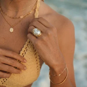 Woman wearing layered 18K gold baroque pearl ring with yellow crochet top at seaside, styled with stacking rings