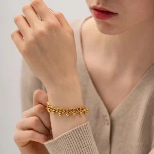 Gold beaded chain bracelet worn on female wrist close-up front view showing snug fit and dangling bead accents with beige knit cardigan styling