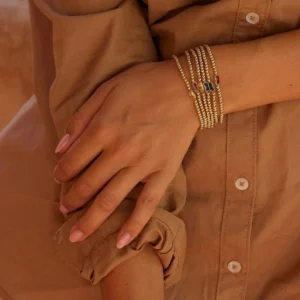 Stacked 18K gold birthstone bracelets worn on wrist with multiple colored baguette gemstones, layered gold beaded chains styling