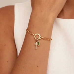 Green birthstone cross charm bracelet worn on wrist, close-up detail of pavé cross and baguette gem with gold paperclip chain