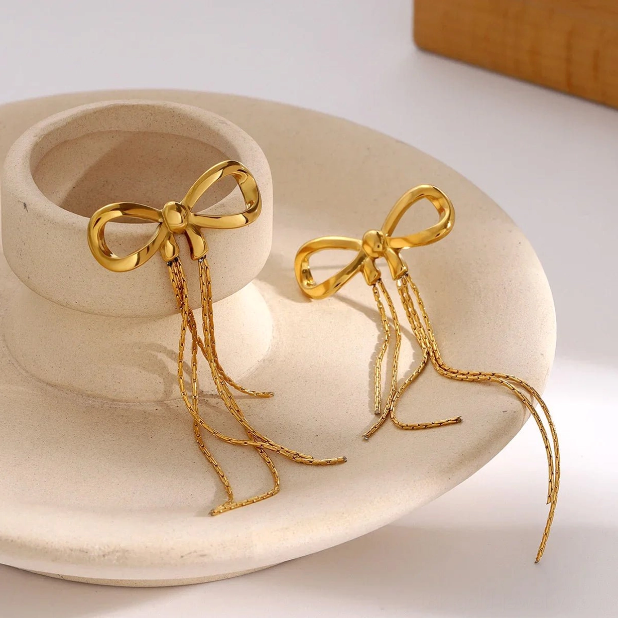 Gold bow stud earrings flat lay on ceramic background, polished 18K gold with chain tassels visible