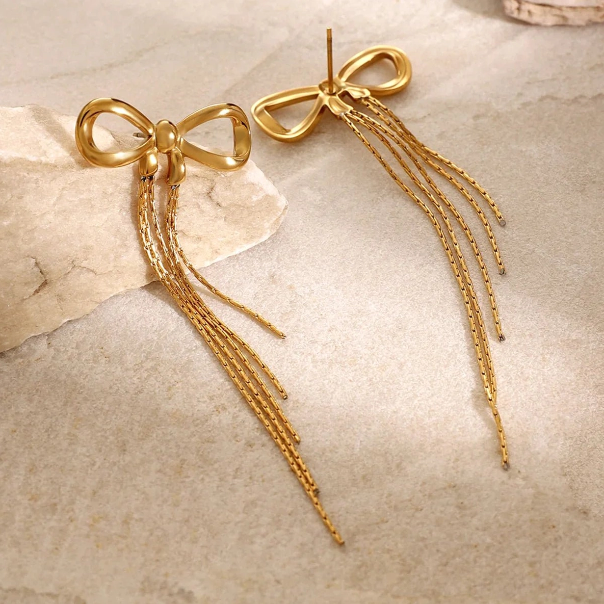 Top-down view of 18K gold bow tassel earrings showing ear posts and polished gold finish on stone texture