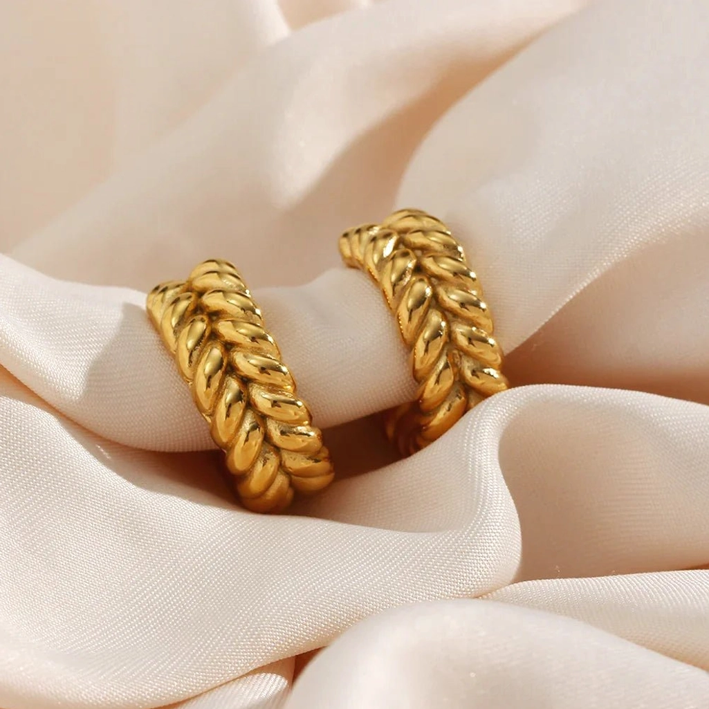 Oblique flat lay of 18K gold braided wheat hoop earrings on beige satin fabric highlighting texture and golden warmth