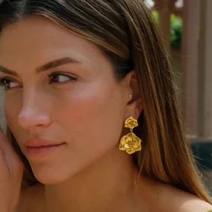 Woman wearing 18K gold camellia drop earrings on left ear outdoor portrait showing warm gold tone and elegant styling on long brown hair