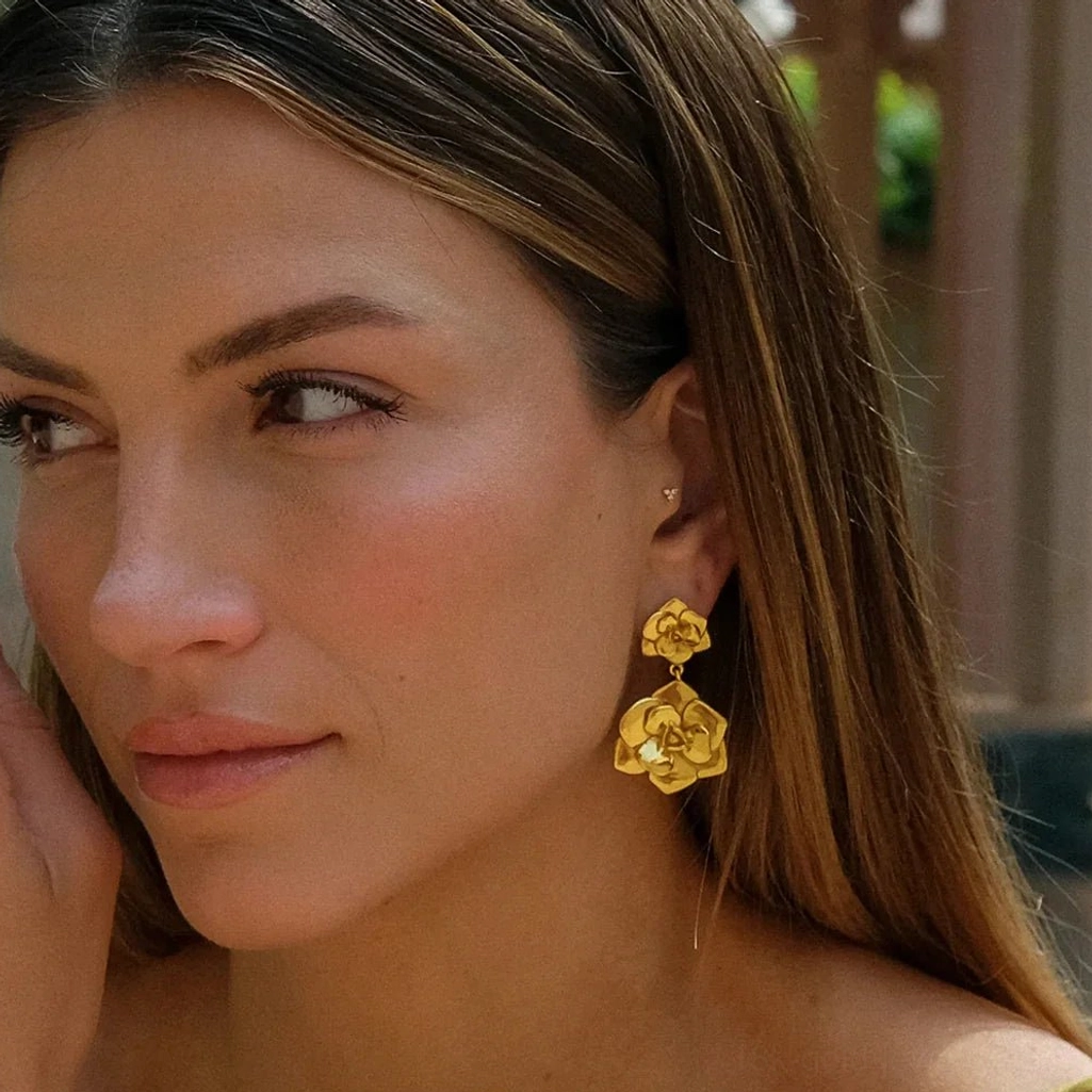 Woman wearing 18K gold camellia drop earrings on left ear outdoor portrait showing warm gold tone and elegant styling on long brown hair
