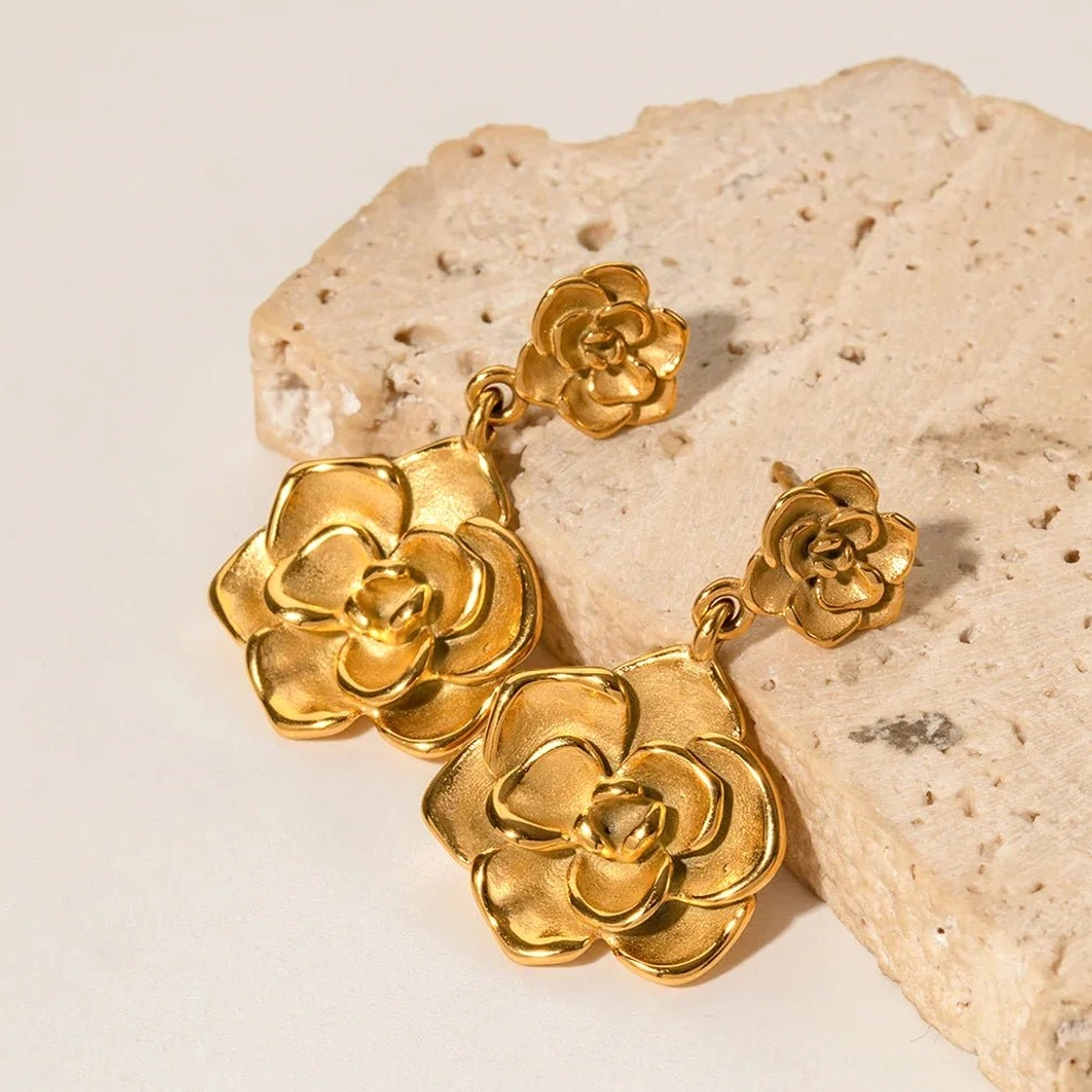 Angled flat lay of 18K gold camellia drop earrings resting on rough stone slab symmetrical double flower design metallic gold surface