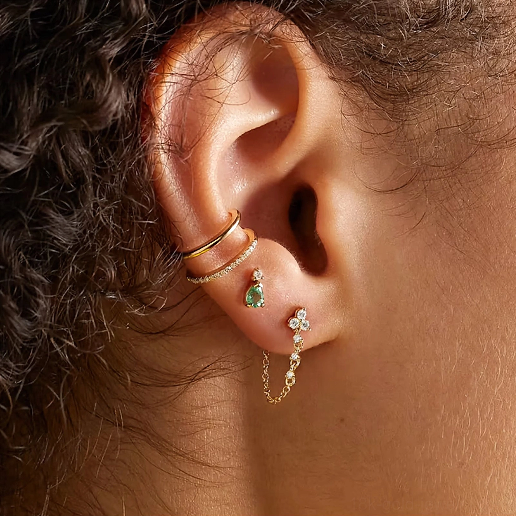 Gold chain stud earrings worn on ear showing layered piercing stack with green gem studs