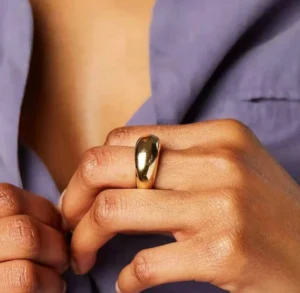 18K gold chunky dome ring worn on hand, close-up eye-level shot, polished gold smooth band, dark-skinned model wearing purple top