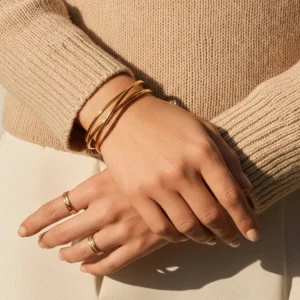 18K gold crisscross cuff bracelet worn on wrist with beige knit sweater and layered gold stack rings, close-up side view showing warm gold tone and multi-band twisted design