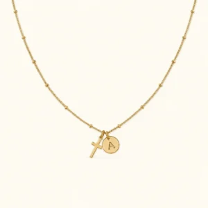 18K gold cross initial necklace flat lay showing beaded satellite chain and personalized hand-stamped coin disc on white background
