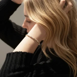 Slim dainty 18K gold CZ bracelet worn on female wrist with blonde hair and black knit sweater, close-up side view showing delicate sparkle