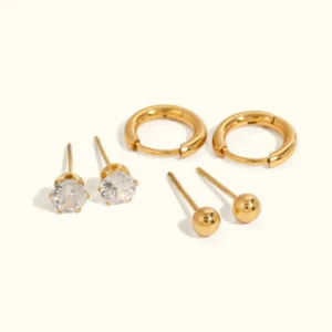 Flat lay studio shot of complete 18K gold earring bundle set on off-white background, displaying all three earring styles including gold studs, CZ diamond studs, and huggie hoops