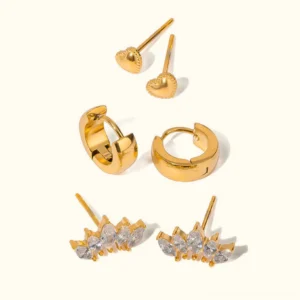 Complete 18K gold earring collection flat lay on plain background, showing all four styles together: heart studs, huggie hoops, and crystal crown studs in one curated set