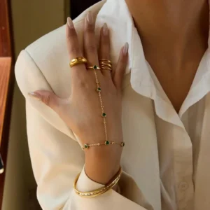 emerald heart hand chain jewelry worn on model wrist close-up view with stacked gold rings and white blazer