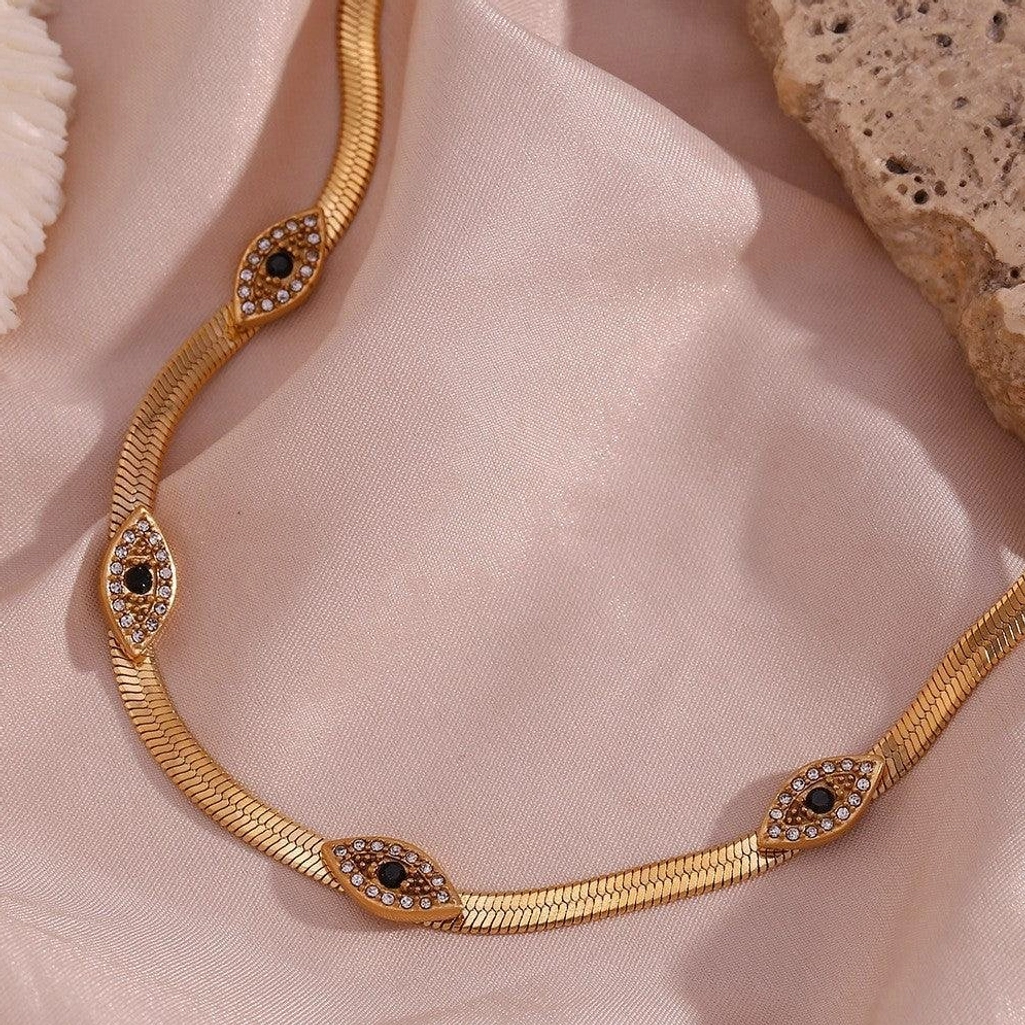 Close-up detail of 18K gold evil eye herringbone necklace on satin fabric highlighting polished chain and crystal-trimmed evil eye charms