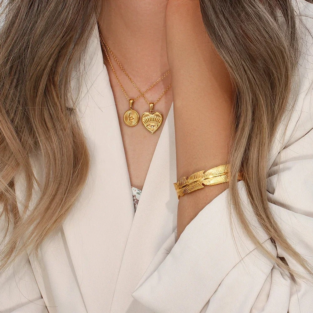 gold feather cuff bracelet stacked with layered heart pendant necklaces on female wrist and neck white blazer