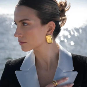18K gold hammered rectangular stud earrings worn on female model's earlobe, side profile view, seaside outdoor setting demonstrating everyday wear