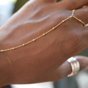 Hand chain jewelry close-up side view worn by dark-skinned model showing gold beaded satellite chain connecting hand and finger