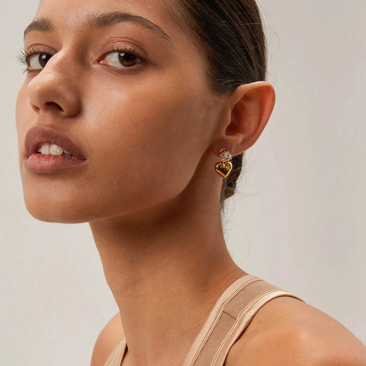 18K gold heart drop earring fitted on earlobe, three-quarter close-up angle on model with neat hairstyle and natural makeup, showing sparkle and craftsmanship