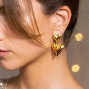 Gold double heart drop earrings worn on ear, close-up side portrait showing glossy metallic finish and elegant dangle