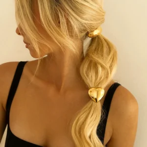 18K gold heart hair cuff worn on blonde model's sleek ponytail, left side profile view showing polished metallic charm detail