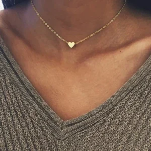 Gold heart necklace worn on model at collarbone, close-up upper body view, woman wearing gray knit top, tiny heart pendant with slim gold chain